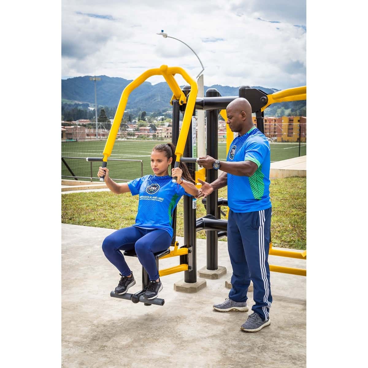Jalón de espalda doble de TMGYM en un parque con montañas al fondo, con una usuaria entrenando y otra persona a su lado