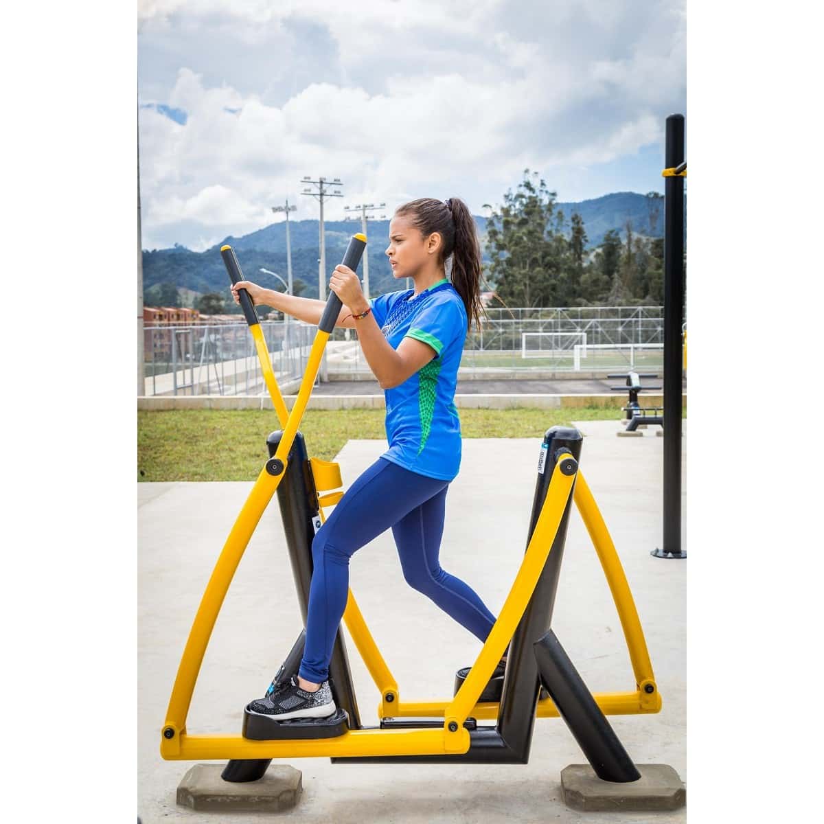 Mujer entrenando en el Sky de TMGYM, equipo de cardio amarillo y negro en un parque con montañas al fondo