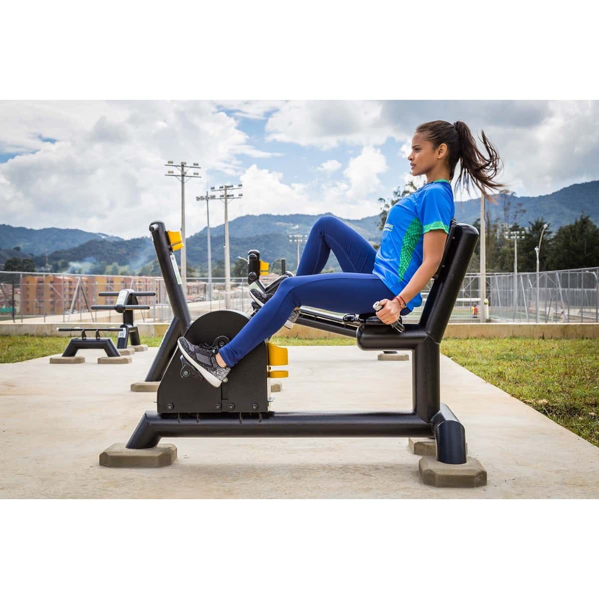 Mujer pedaleando en la bicicleta recumbent de TMGYM, instalada en un parque con montañas al fondo