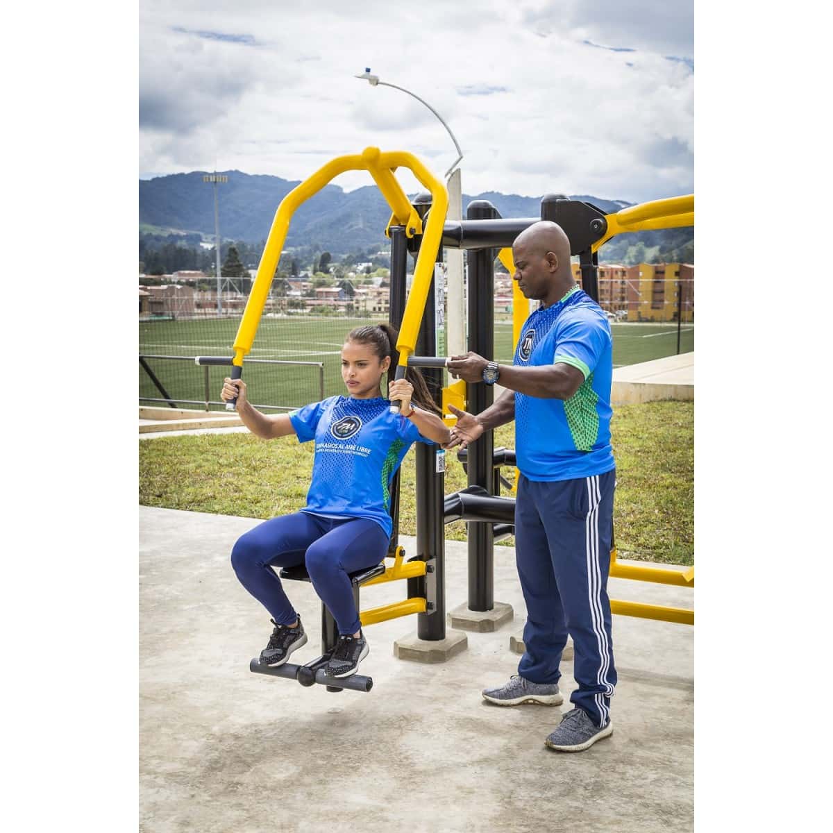 Prensa de pecho doble de TMGYM en un parque con montañas al fondo, con una usuaria entrenando y otra persona a su lado