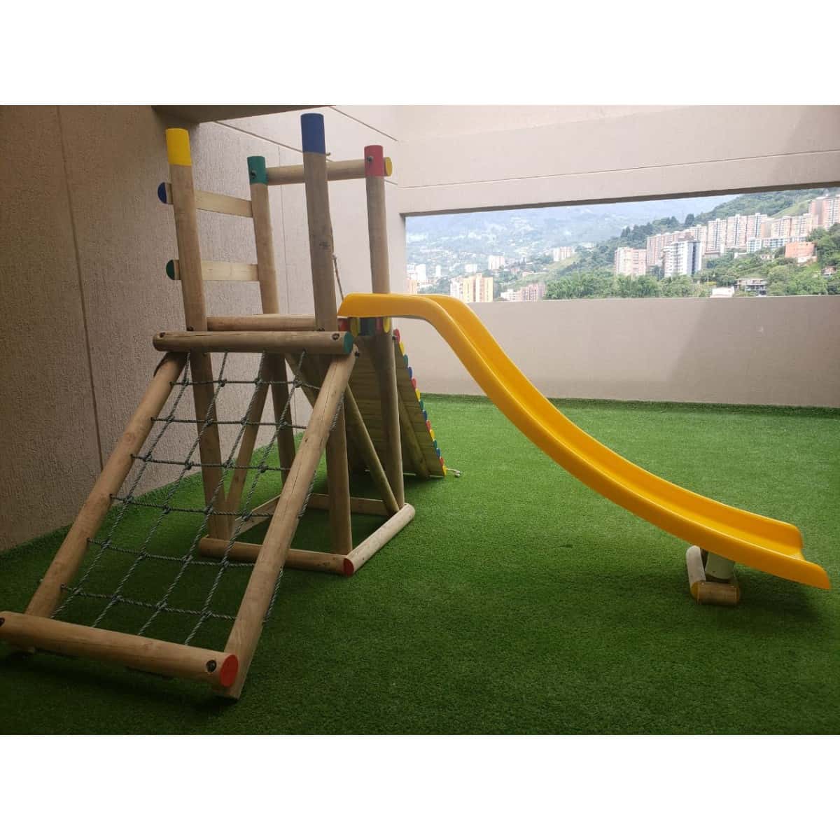 Parque Tayrona de madera con red de trepa y deslizadero amarillo sobre grama sintética, junto a una ventana con vista a la ciudad