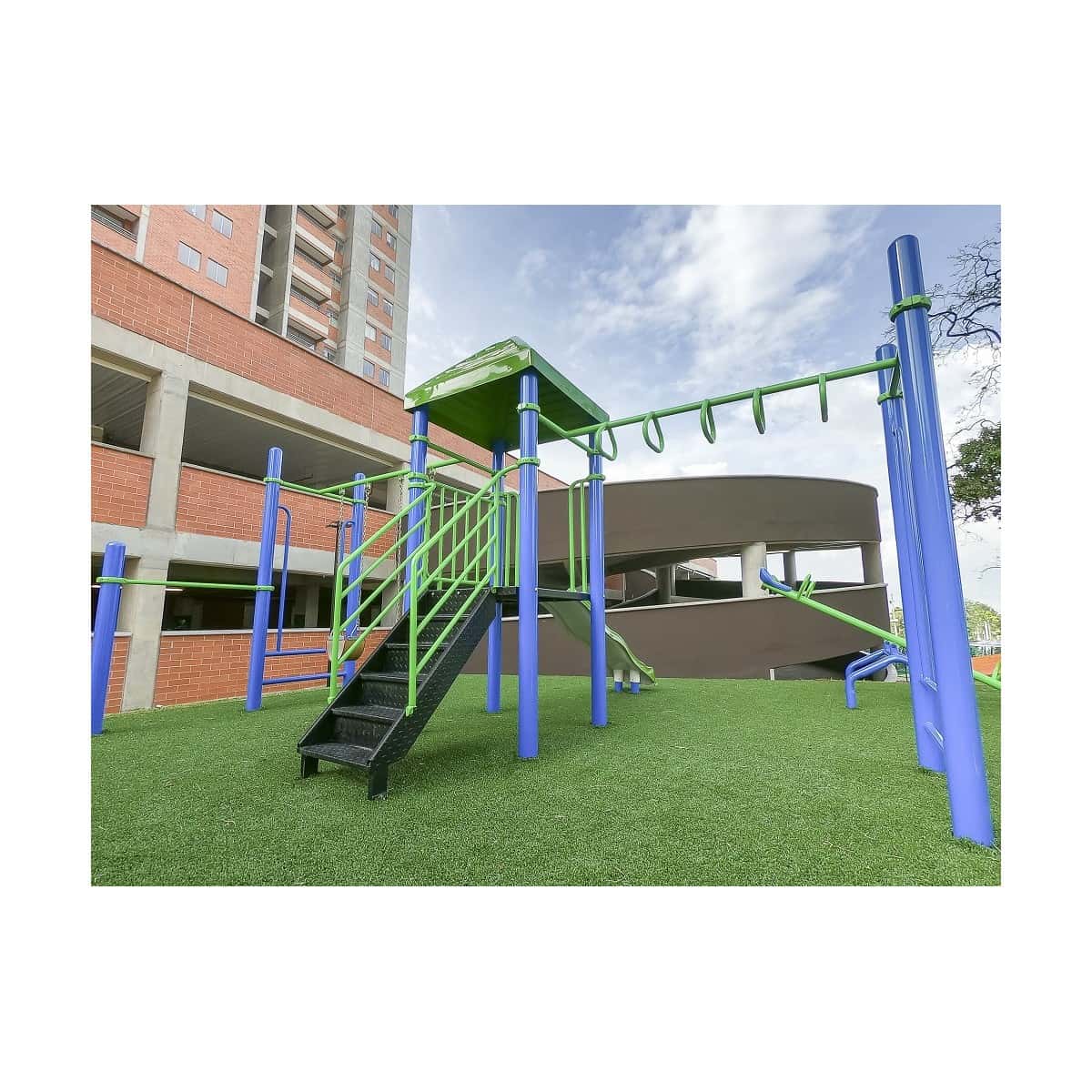 Parque infantil recreativo con torre de techo verde, escalera y pasamanos sobre grama sintética, junto a un edificio de apartamentos