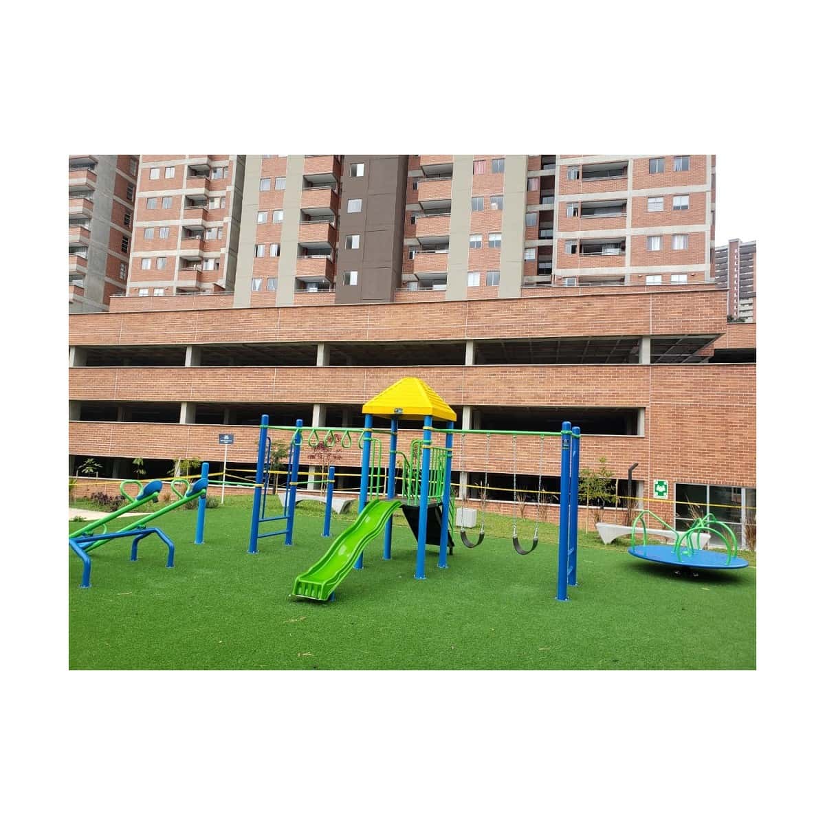 Parque infantil recreativo con columpios, balancín y carrusel sobre grama sintética frente a un edificio de apartamentos