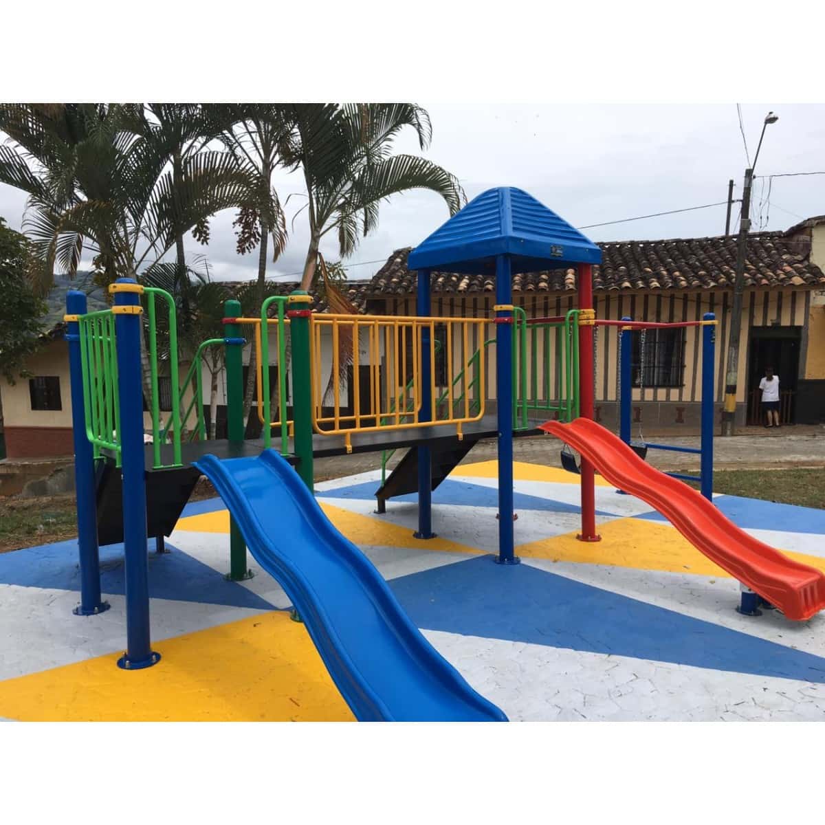 Parque infantil con techo azul, deslizaderos y columpios frente a una vivienda