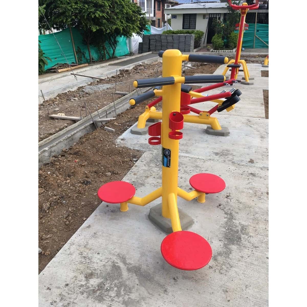 Twister de cintura de TMGYM, equipo amarillo con discos rojos instalado sobre concreto junto a una obra de parque