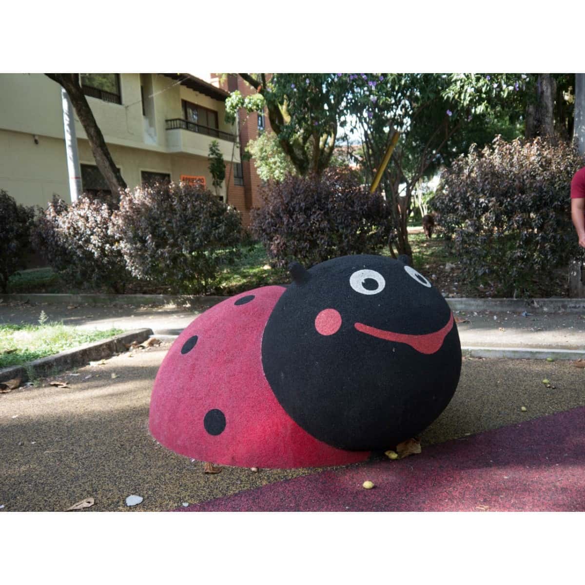 Figura 3D de mariquita instalada en un parque con árboles y edificios al fondo