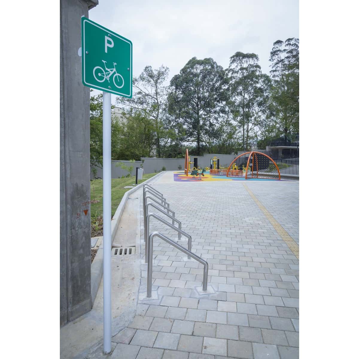 Parquearos metálicos para bicicletas en una plaza con piso adoquinado y un parque al fondo