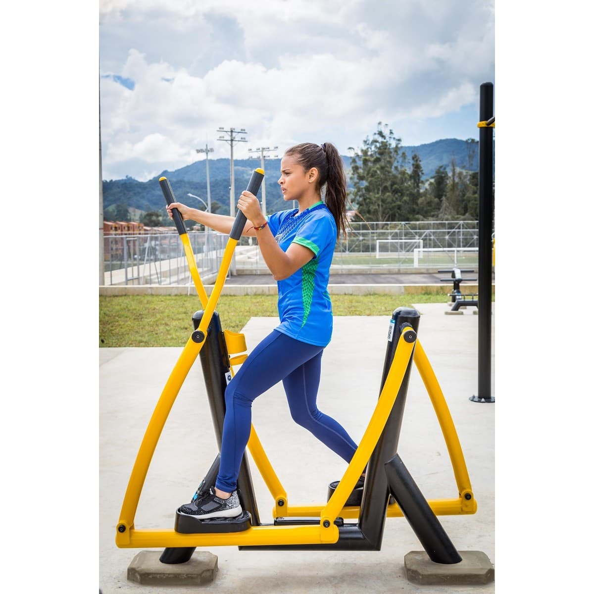 Mujer entrenando en el Sky de TMGYM, equipo de cardio amarillo y negro en un parque con montañas al fondo