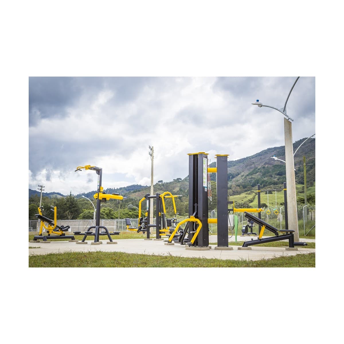 Estación multifuncional de TMGYM junto a otros equipos de gimnasio al aire libre en un parque con montañas al fondo
