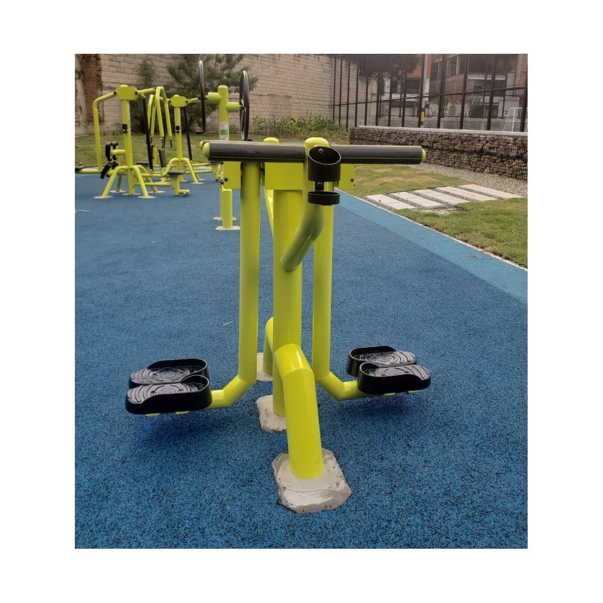 Péndulo de TMGYM, equipo amarillo con pedales y agarres sobre piso azul en un gimnasio al aire libre