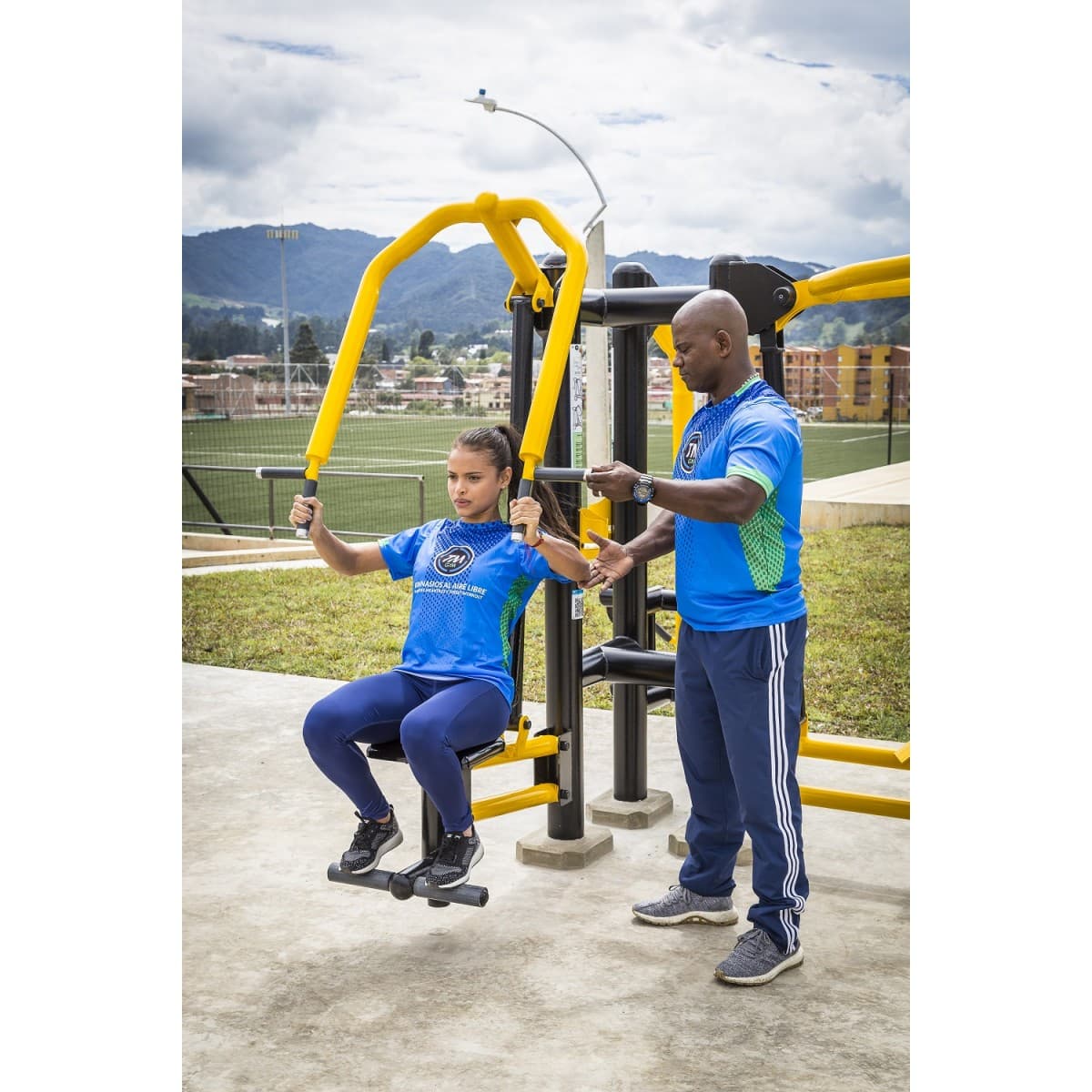 Prensa de pecho doble de TMGYM en un parque con montañas al fondo, con una usuaria entrenando y otra persona a su lado