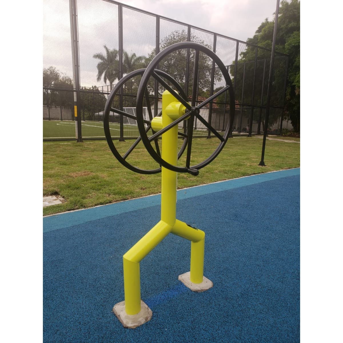 Rueda de hombros de TMGYM, equipo con rueda negra y estructura amarilla sobre piso azul en un parque