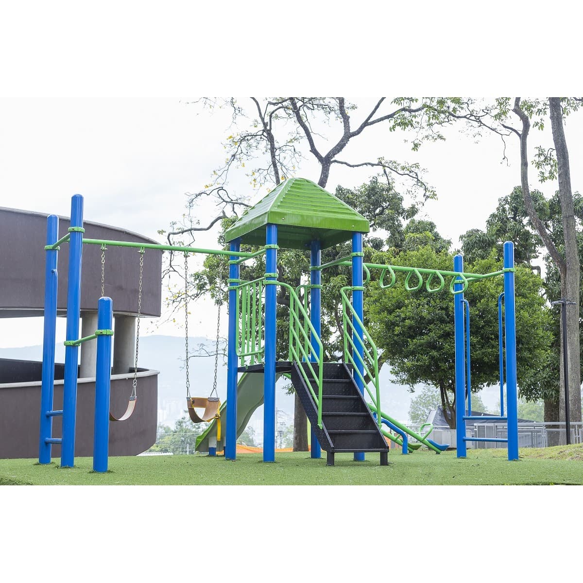 Parque infantil recreativo metálico con techo verde, estructura azul, escalera y pasamanos en un parque con árboles