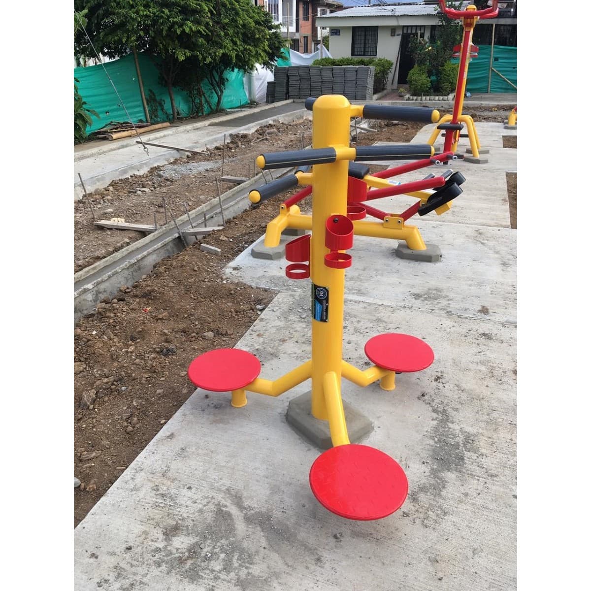 Twister de cintura de TMGYM, equipo amarillo con discos rojos instalado sobre concreto junto a una obra de parque